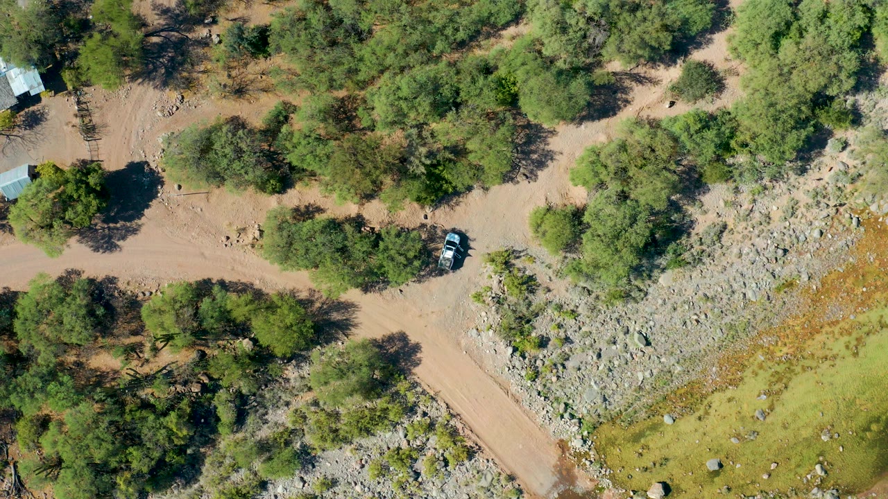 Overhead frame from San Javier, Baja California Sur showing a light-colored car on a tan dirt road beside green trees, rocky terrain, and no visible people.