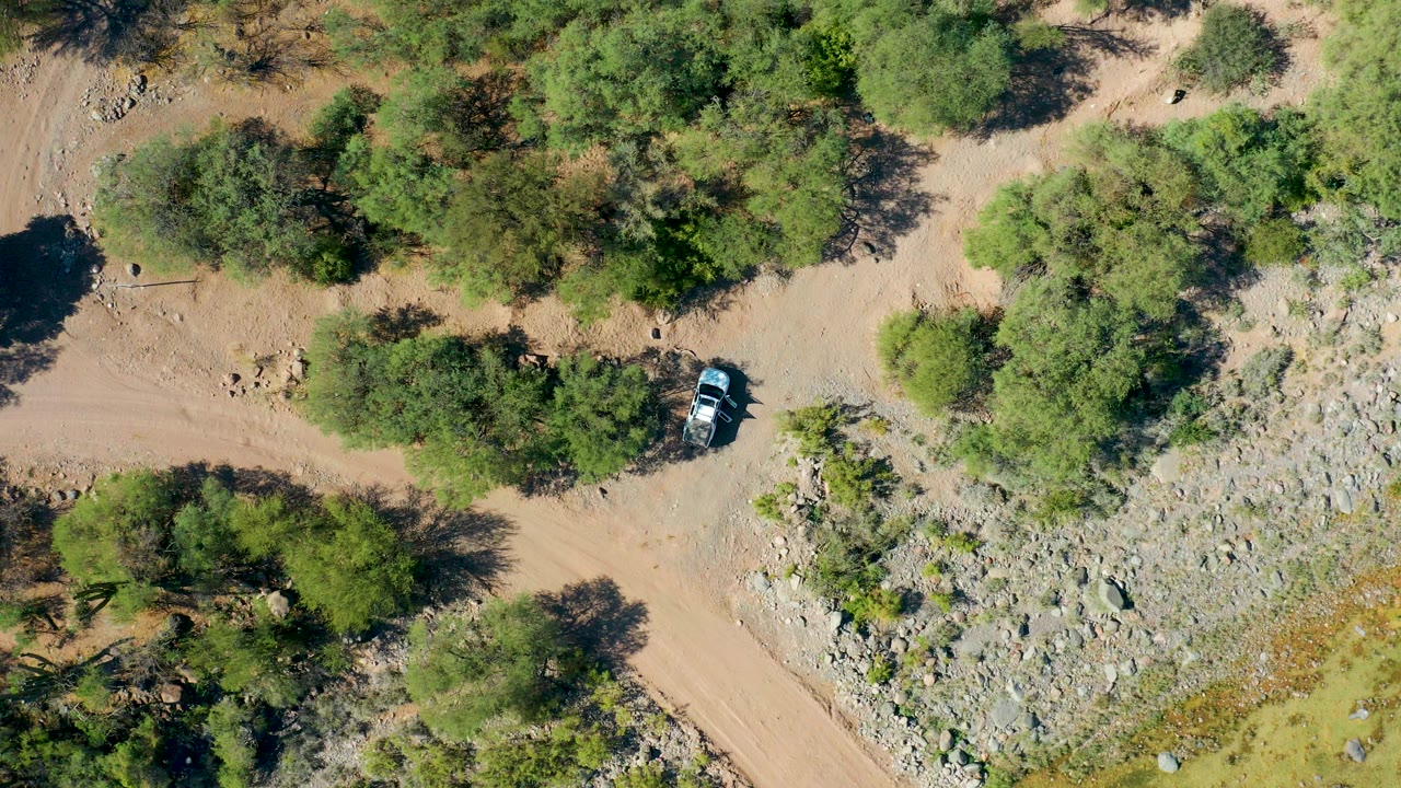 Overhead frame in San Javier, Baja California Sur showing a light-colored car on a tan dirt road beside green trees, rocky ground, and no visible people.