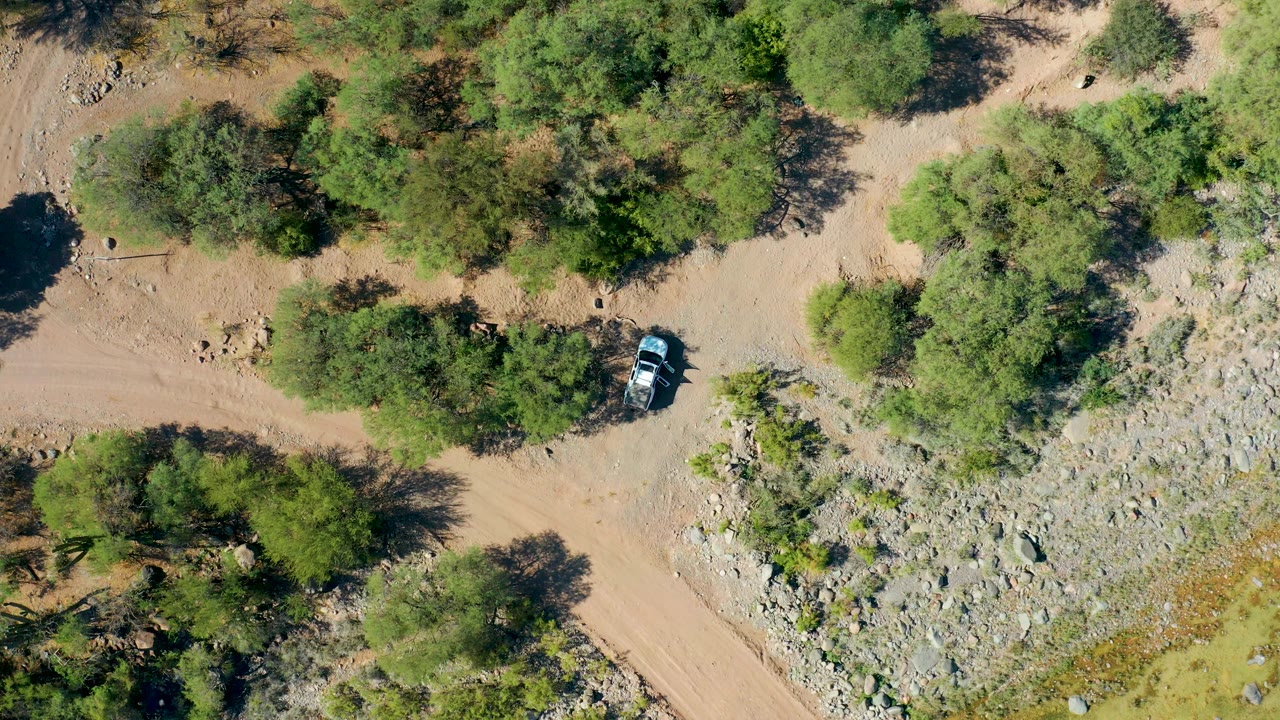 Drone-like overhead frame in San Javier, Baja California Sur showing the car near the edge of a tree cluster, with a broad rocky wash to one side and no visible people.