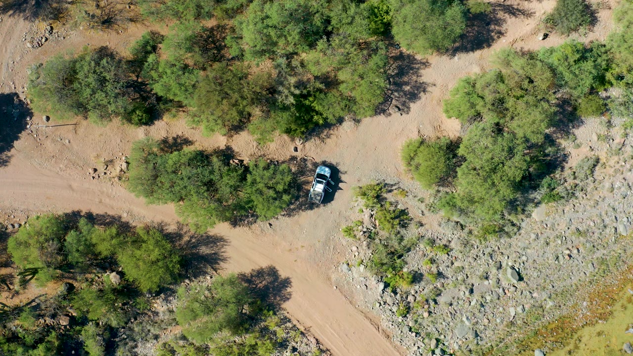 Overhead San Javier, Baja California Sur frame with the car lower in the road corridor, surrounded by tan dirt, green trees, dark shadows, and scattered rocks, with no visible people.