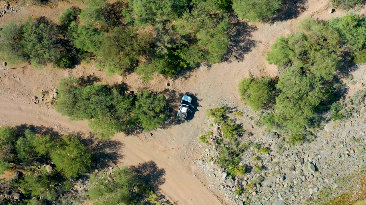 Aerial frame in San Javier, Baja California Sur with the vehicle advancing along the dirt road near thicker tree cover and a pale rocky wash, with no visible people.