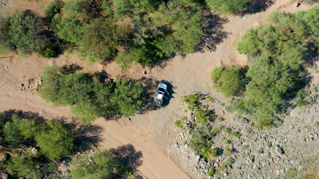 Overhead view from San Javier, Baja California Sur with the vehicle near the edge of a rocky drainage area, tan road, green shrubs, and no visible people.