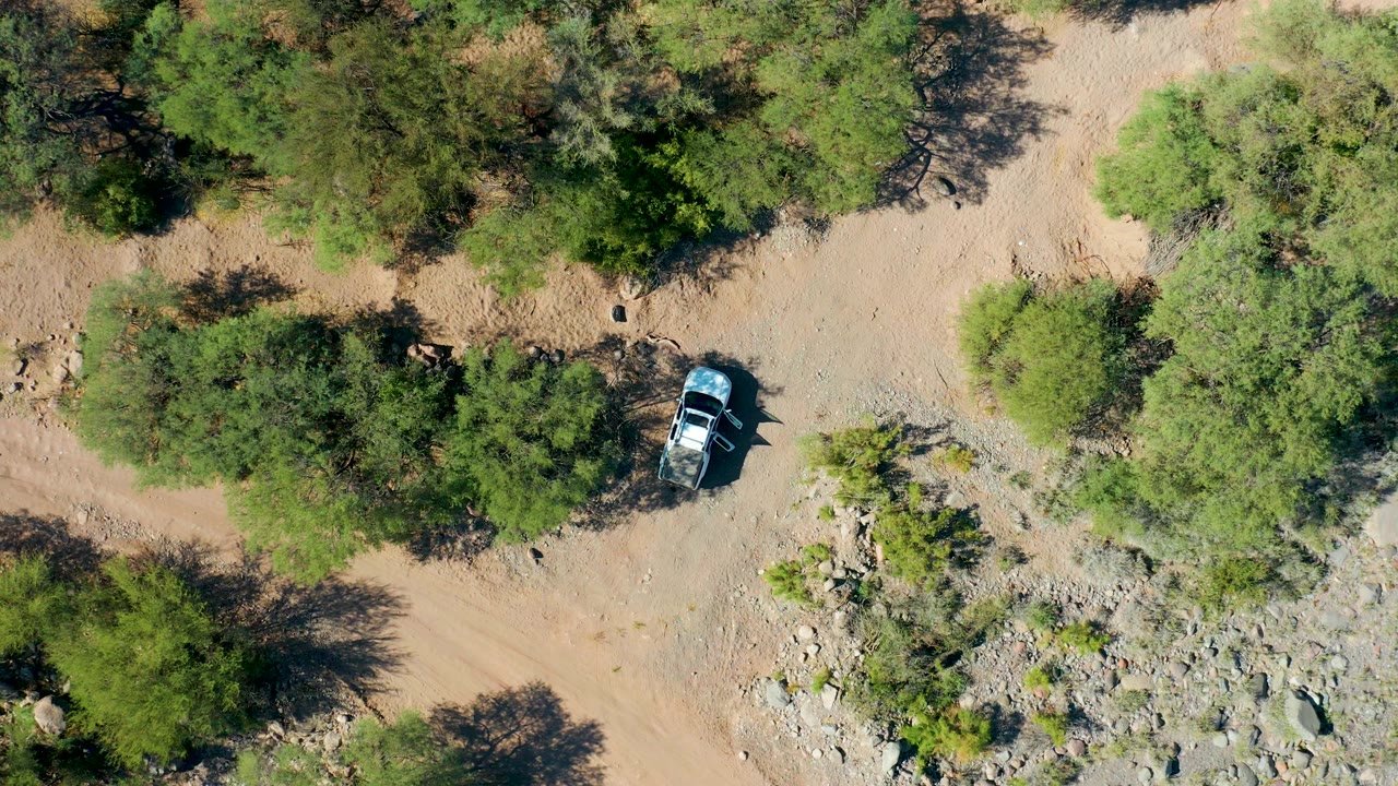 Overhead San Javier frame with the car slightly farther along the tan trail, bordered by dense green tree cover, scattered rocks, and no visible people.