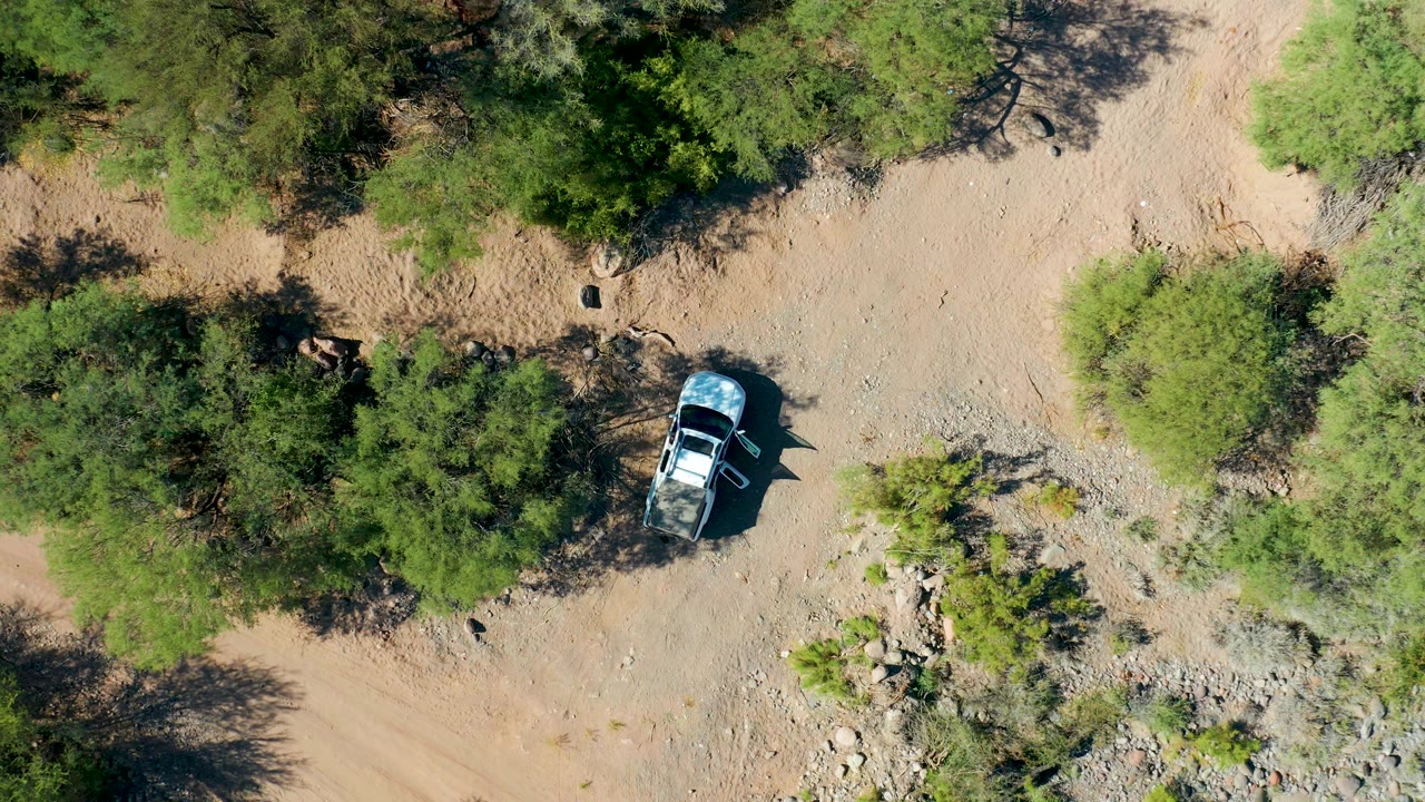 Aerial view over San Javier, Baja California Sur, with the vehicle slightly farther along the pale dirt route near dense green tree cover and no visible people.