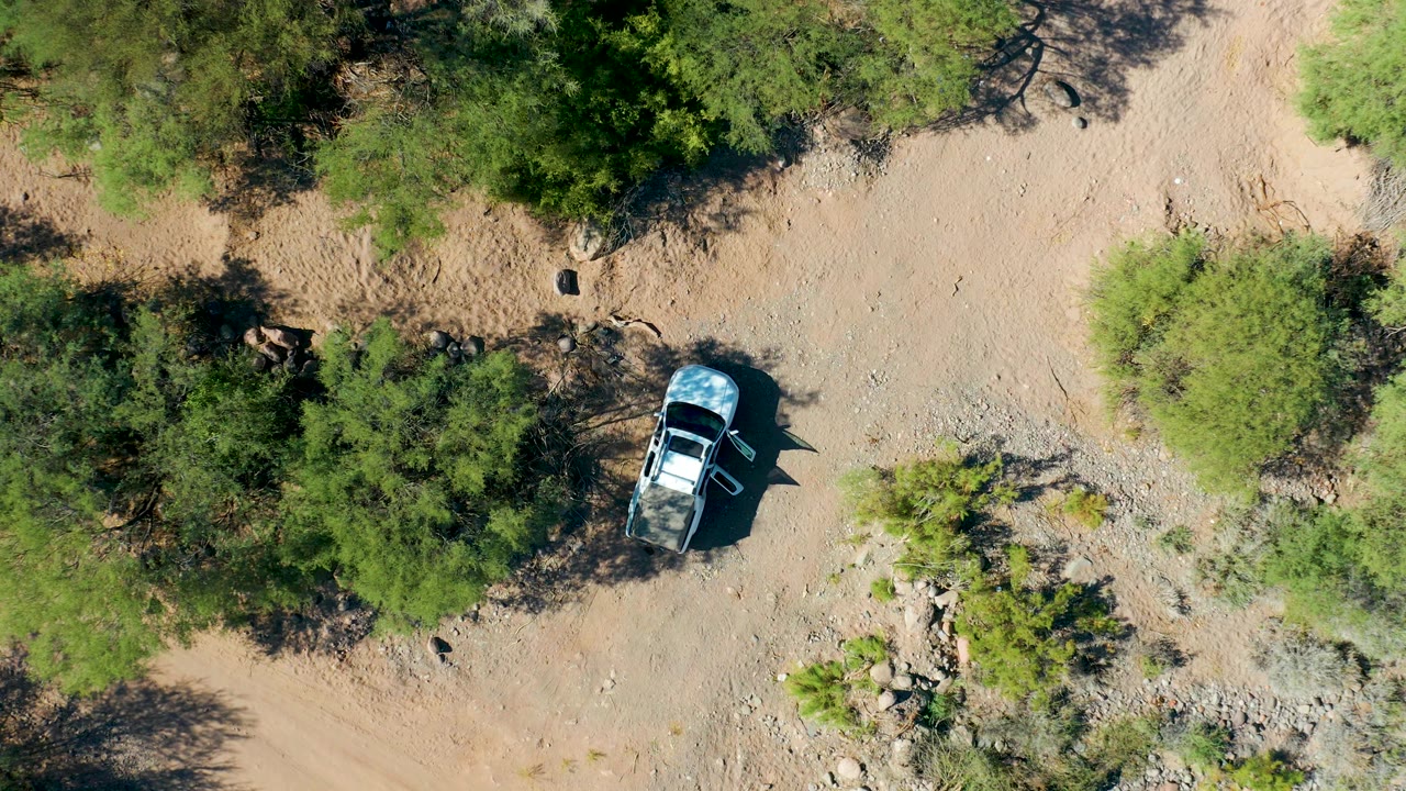 Aerial frame over San Javier, Baja California Sur, with the vehicle partly framed by tree shadows, pale dirt, scrub, rocks, and no visible people.