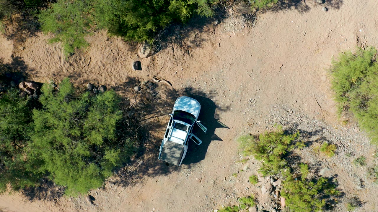 Aerial view in San Javier, Baja California Sur with the white SUV slightly lower in frame, open dry ground above it, green shrubs along the edges, and no visible people.