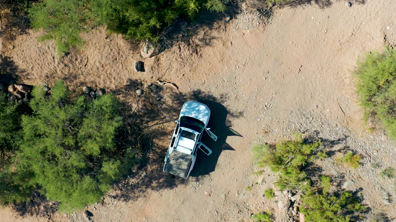 Overhead San Javier frame showing the parked white SUV beside denser green shrubs, dark angular shadows, scattered rocks, and empty arid ground with no visible people.