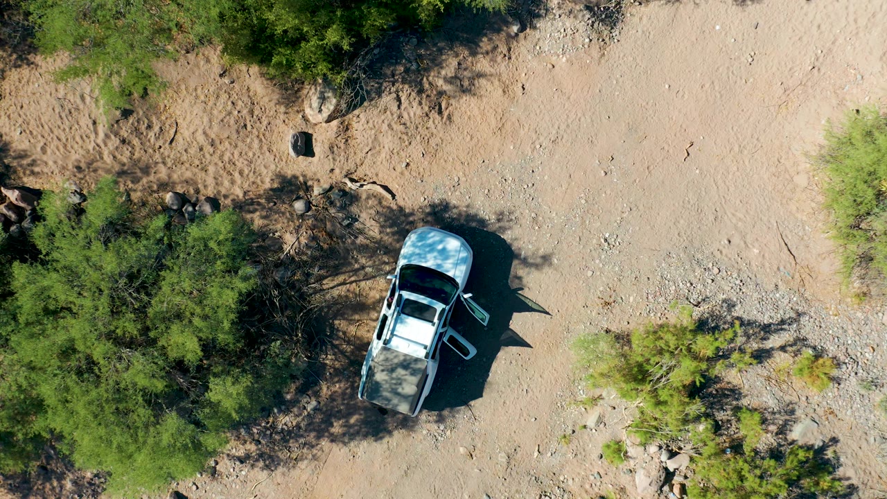 San Javier aerial frame with the white SUV near the lower-left portion of the image, open rocky dirt to the right, green shrubs nearby, and no visible people.