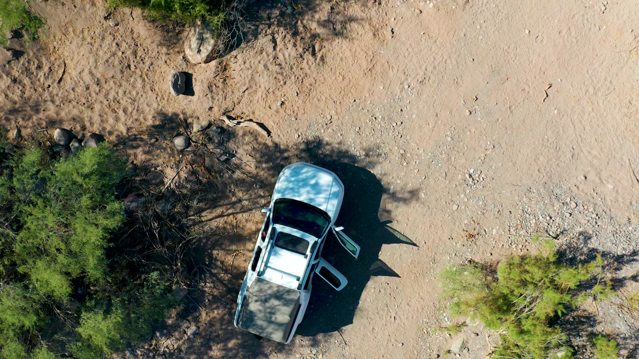 Overhead frame in San Javier, Baja California Sur shows a white SUV parked on pale sandy terrain with one side door open, sparse green trees at the edges, rocks nearby, and no visible people.