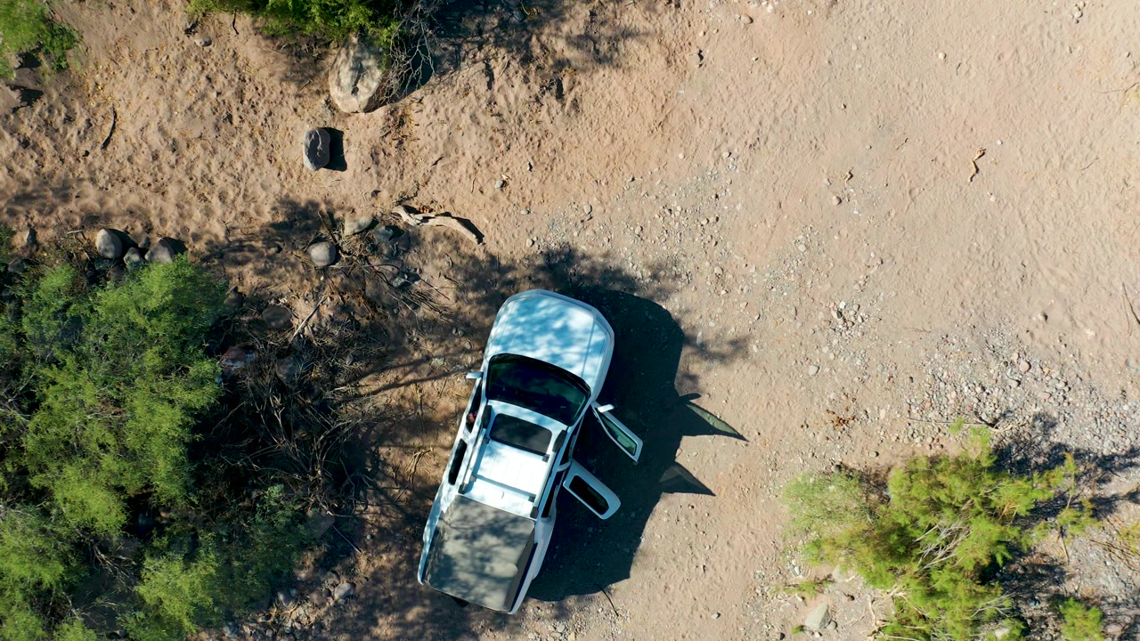 Aerial view in San Javier, Baja California Sur places the white SUV lower in frame with its door open, green desert brush along the left edge, scattered rocks above, and no visible people.