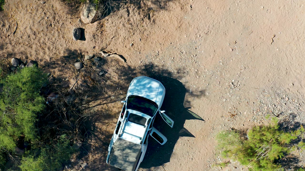 Aerial frame in San Javier, Baja California Sur keeps the white SUV near the lower left with the open door visible, dense tree shadow crossing the sandy surface, scattered stones above, and no visible people.