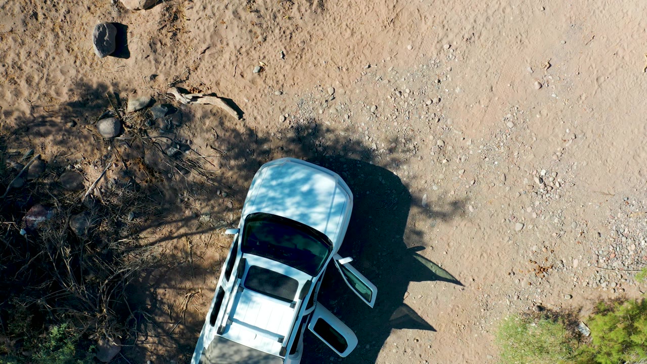 Overhead frame in San Javier, Baja California Sur showing the white car near the lower right of the road, with both the pale gravel surface and dark brush shadows clearly visible and no visible people.