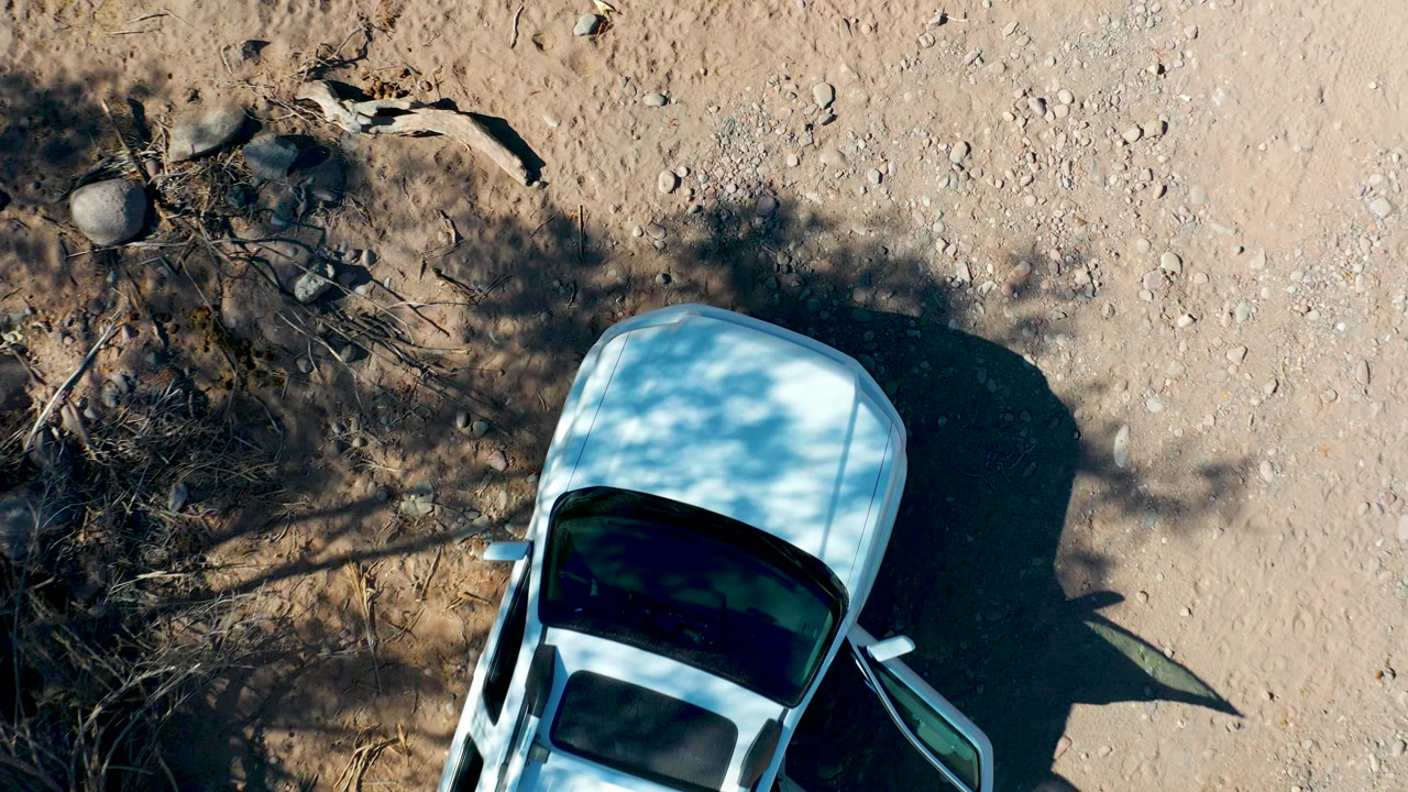Overhead frame in San Javier, Baja California Sur showing the rear portion of a white car at the lower edge, pale gravel, dry branches, tree shadows, and no visible people.