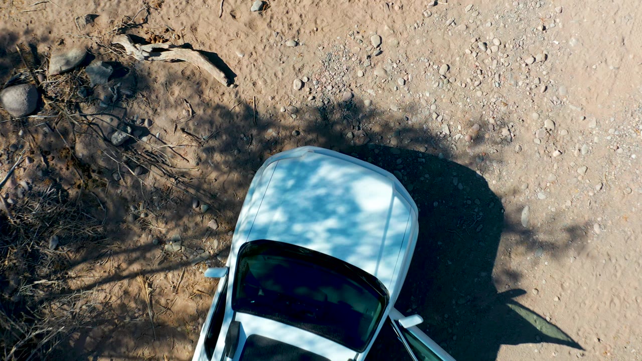 Overhead frame in San Javier, Baja California Sur with the white car cropped at the lower right, a large shadow to the right, dry branches to the left, and no visible people.
