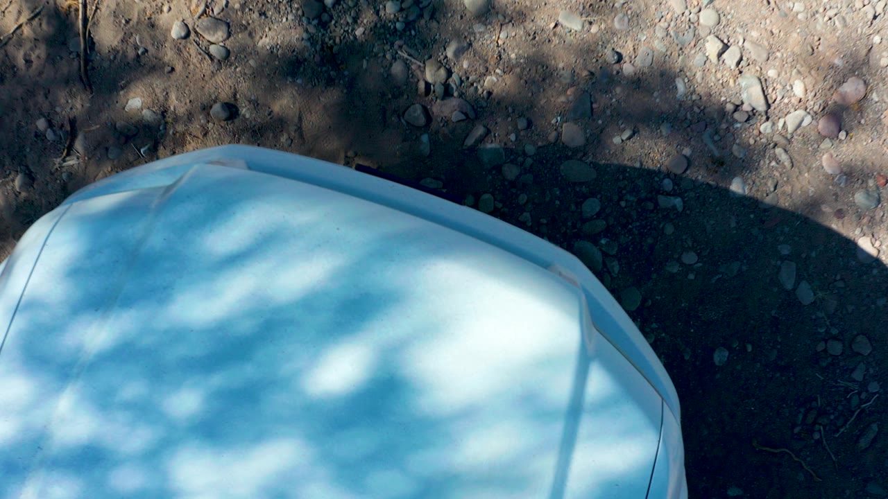 At 0:00 in San Javier, Baja California Sur, a pale vehicle-like hood fills the lower frame beside gravelly ground and tree shadows, with no visible people.