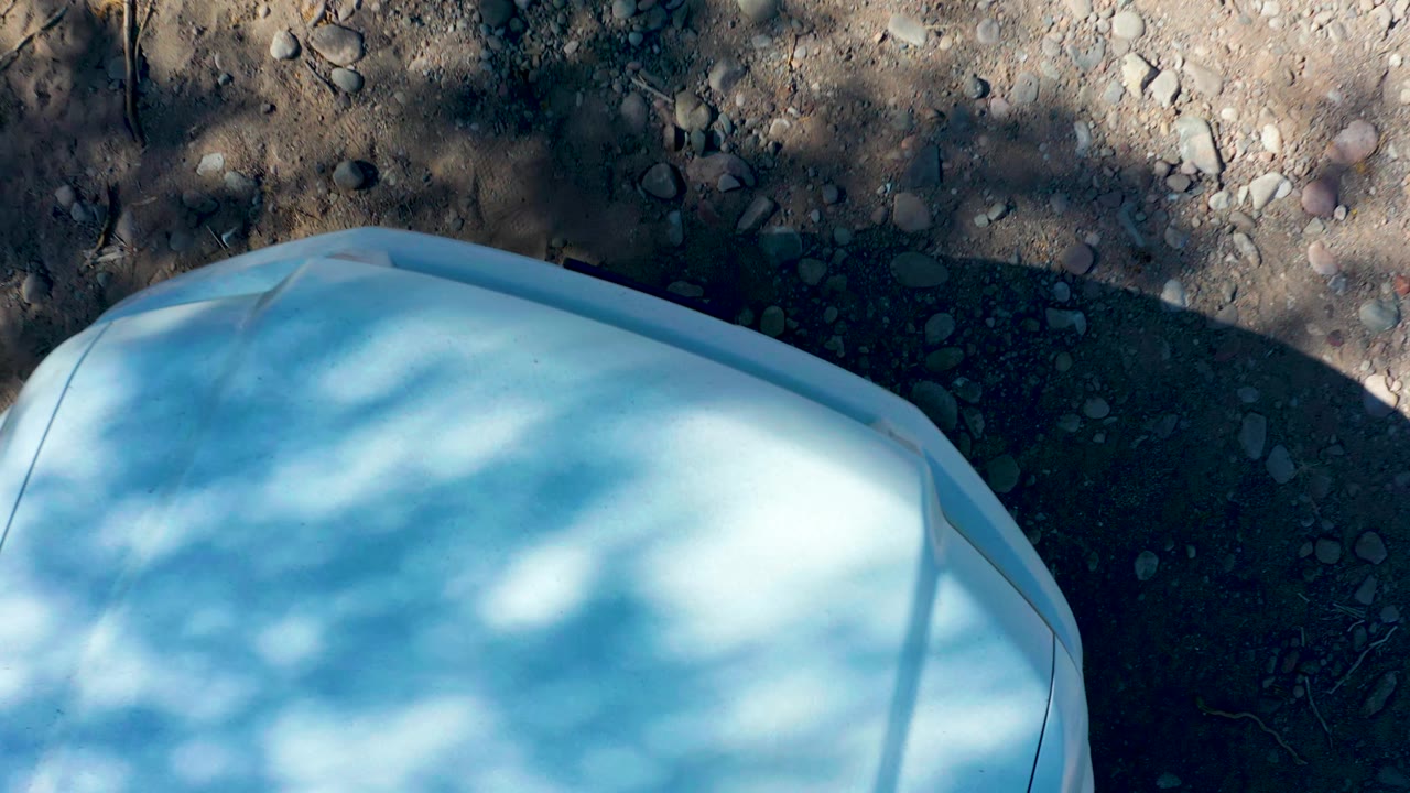 At 0:01 in San Javier, Baja California Sur, the pale hood edge remains low in frame while rocky dirt and broken shade fill the upper area, with no visible people.