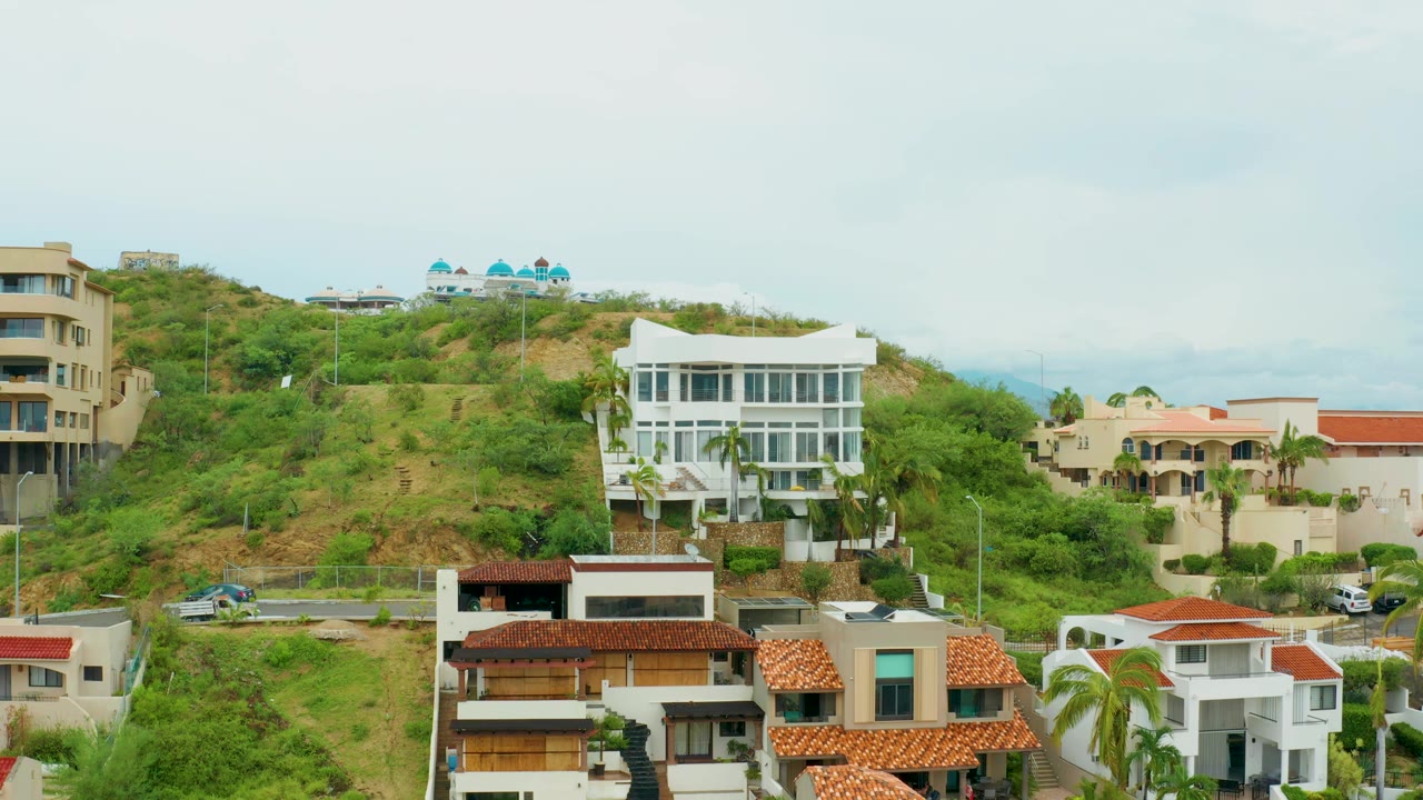 Elevated view of a San Jose del Cabo hillside residential area with green vegetation, white and beige houses, red tile roofs, parked cars, and no visible people.