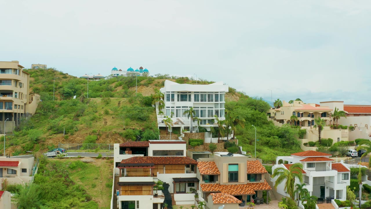 Elevated San Jose del Cabo residential frame showing a white hillside building, beige multi-story homes, red roof tiles, dense greenery, and no visible people.