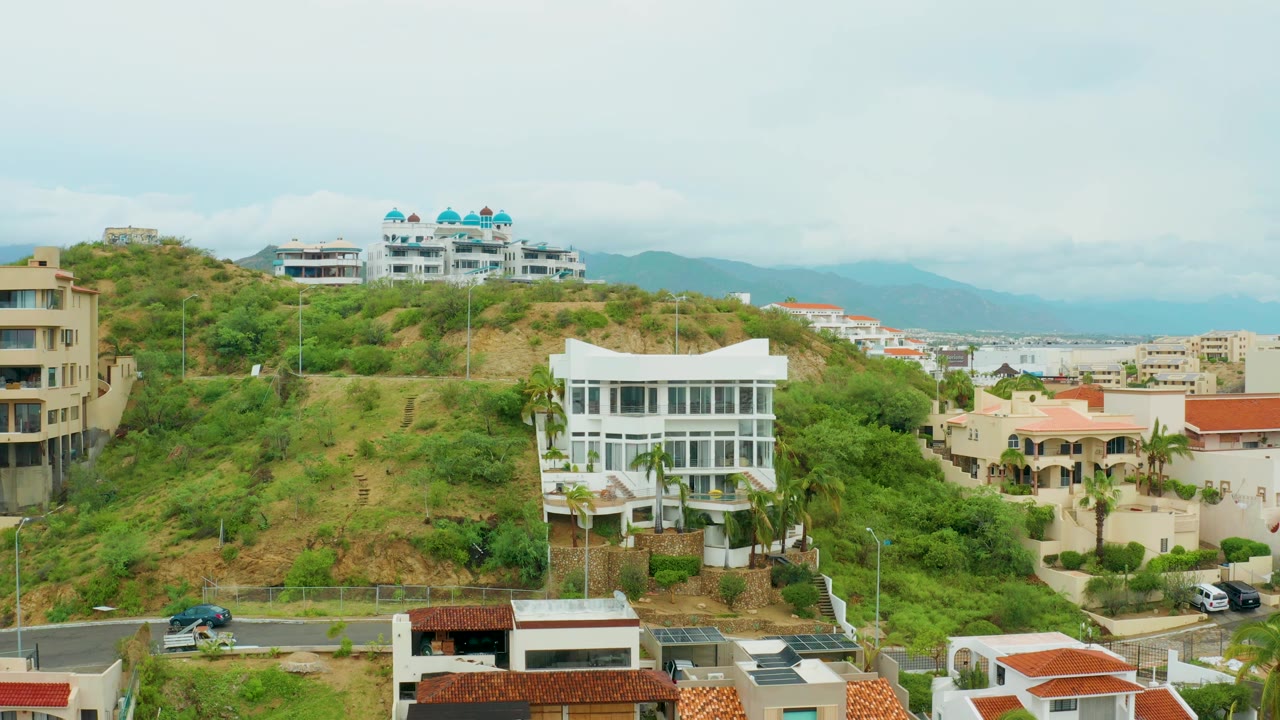 San Jose del Cabo hillside neighborhood with pale residential buildings, dense green vegetation, red roofs, hazy mountains, and no visible people at 0:02.