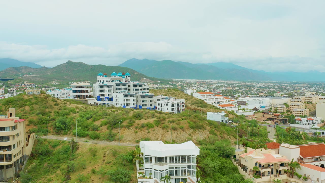 San Jose del Cabo hillside view with a large white residential building on the ridge, lower homes in front, a city plain beyond, mountains, and no visible people.