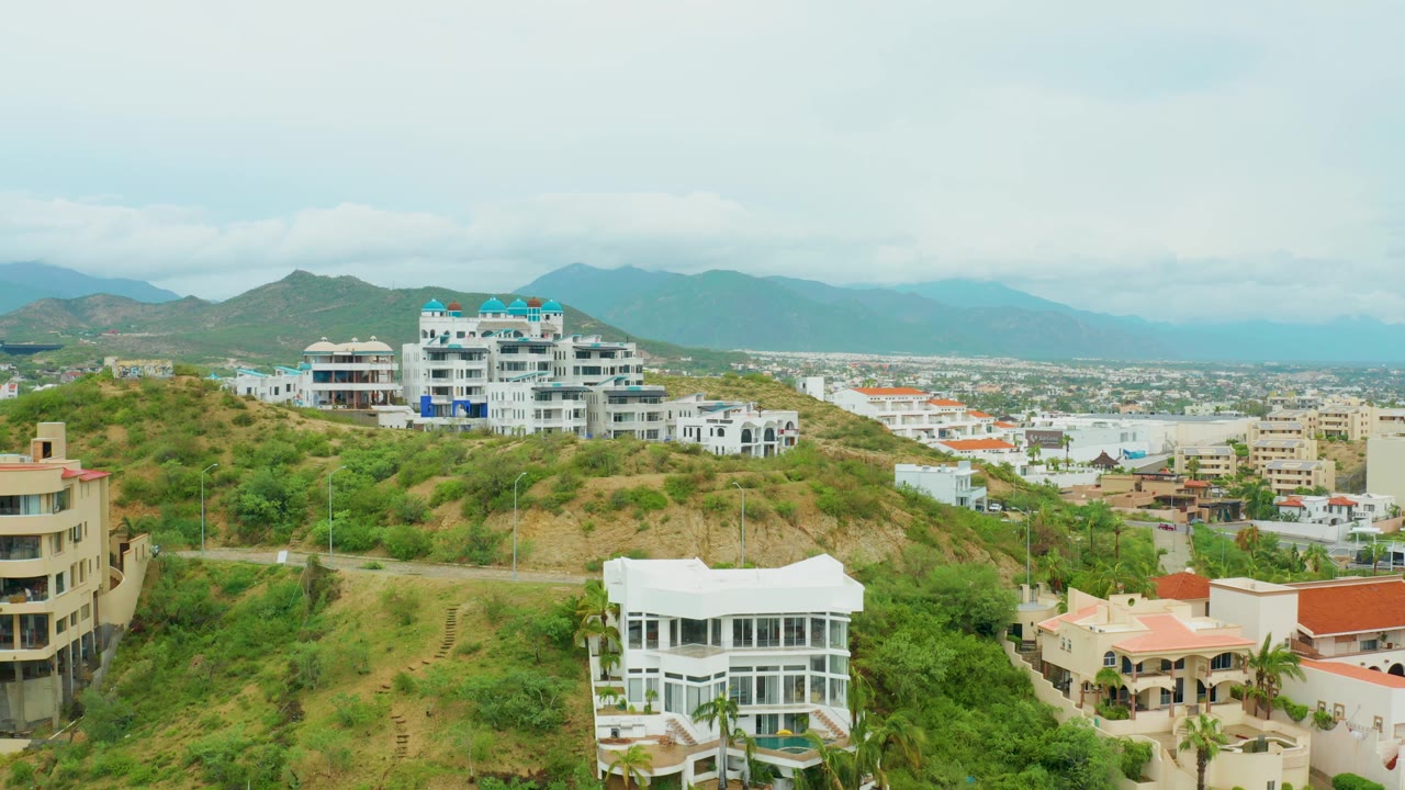 Aerial hillside neighborhood with white homes, vegetation, city, and cloudy mountains.
