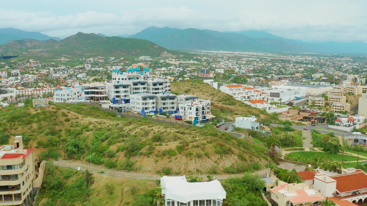 Aerial frame of San Jose del Cabo with white multi-story hillside buildings, a curving road below, broad low-rise development, and no visible people.