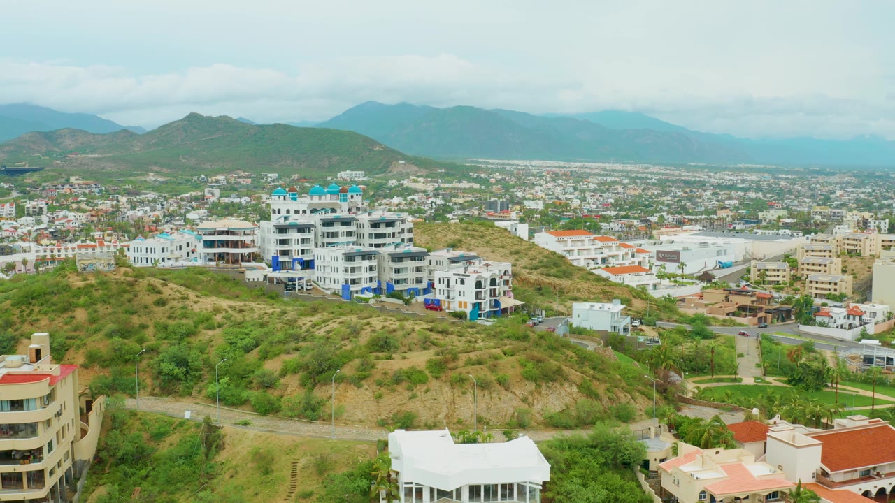 Aerial city view over scrubby hillside, white buildings, road, and mountain backdrop.