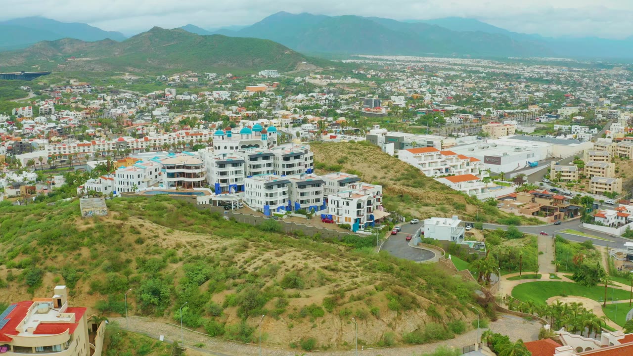 Early frame in San Jose del Cabo, Baja California Sur, with the white residential complex centered, dry hillside foreground, city blocks beyond, and no visible people.
