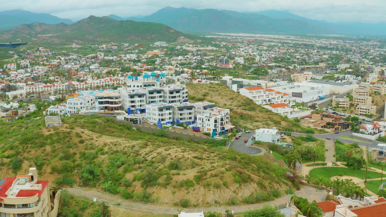 San Jose del Cabo aerial frame showing hillside apartments near the center-left, pale commercial roofs and city streets to the right, mountains behind, and no visible people.