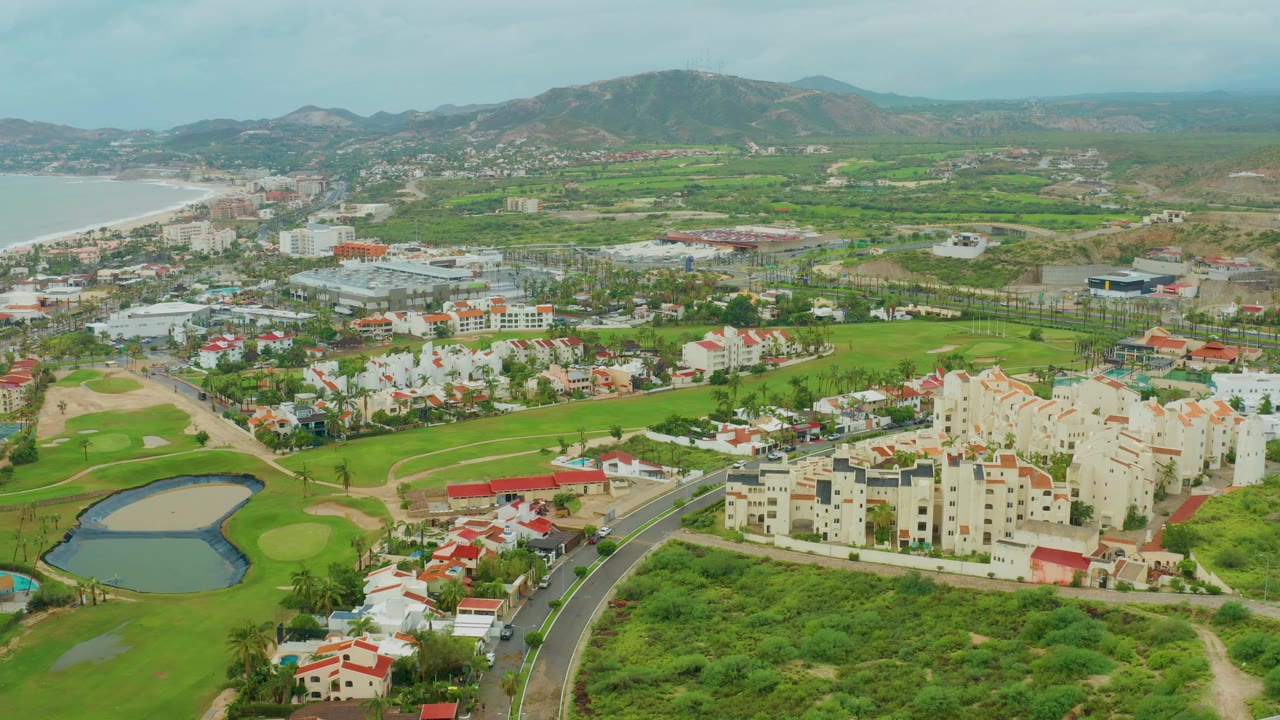 Wide aerial frame over San Jose del Cabo BCS Mexico showing a road curving past a golf course pond, cream residential buildings, distant hills, shoreline, and no visible people.