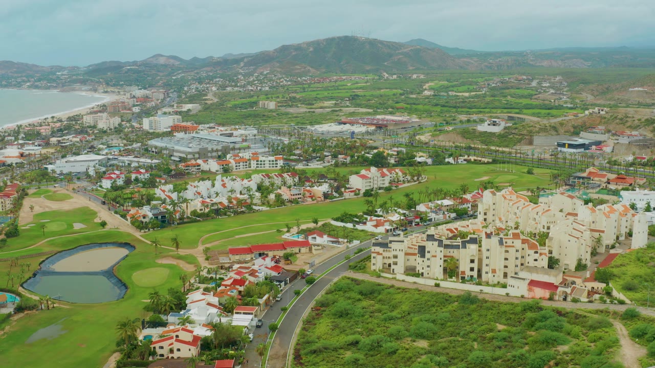 Elevated view of green fairways, a pond, residential buildings, a road, hills, and distant water.