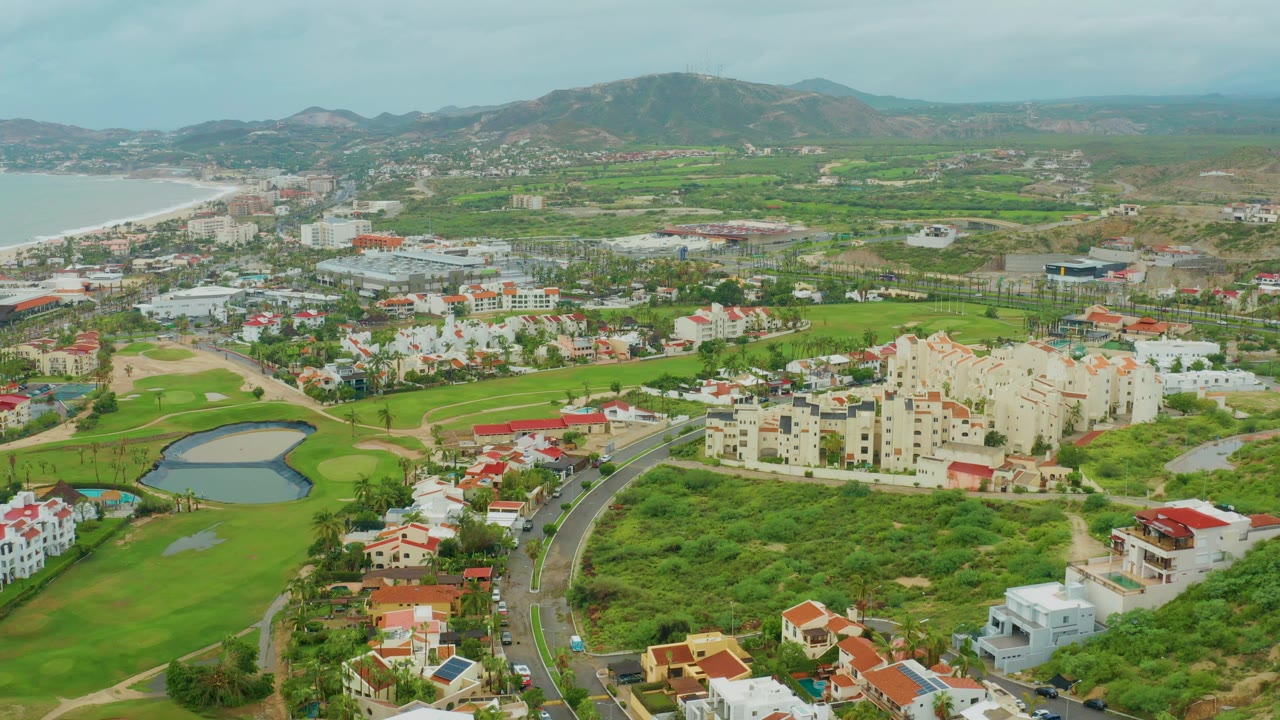 Aerial frame from San Jose del Cabo, Baja California Sur showing a curved road beside a green golf course pond, residential buildings, ocean at left, distant hills, and no visible people.