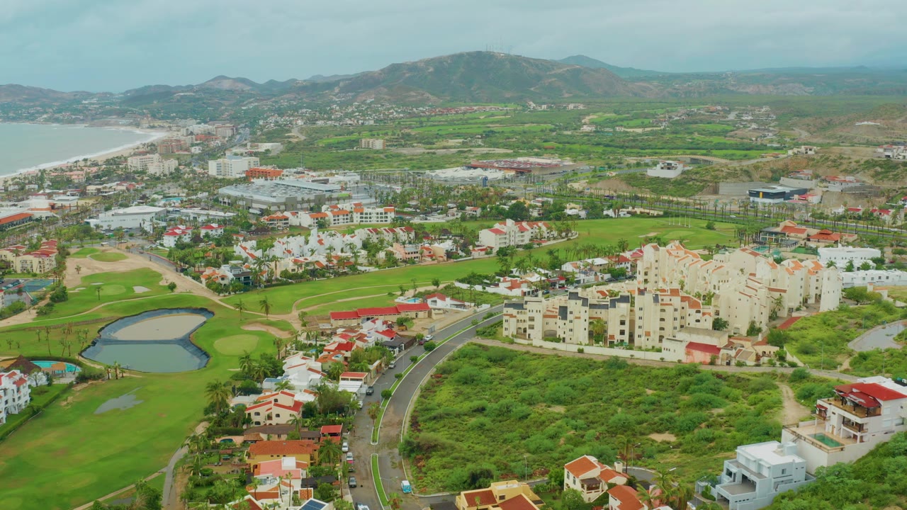 Aerial view of coastal development with golf greens, a pond, traffic, homes, and hills.