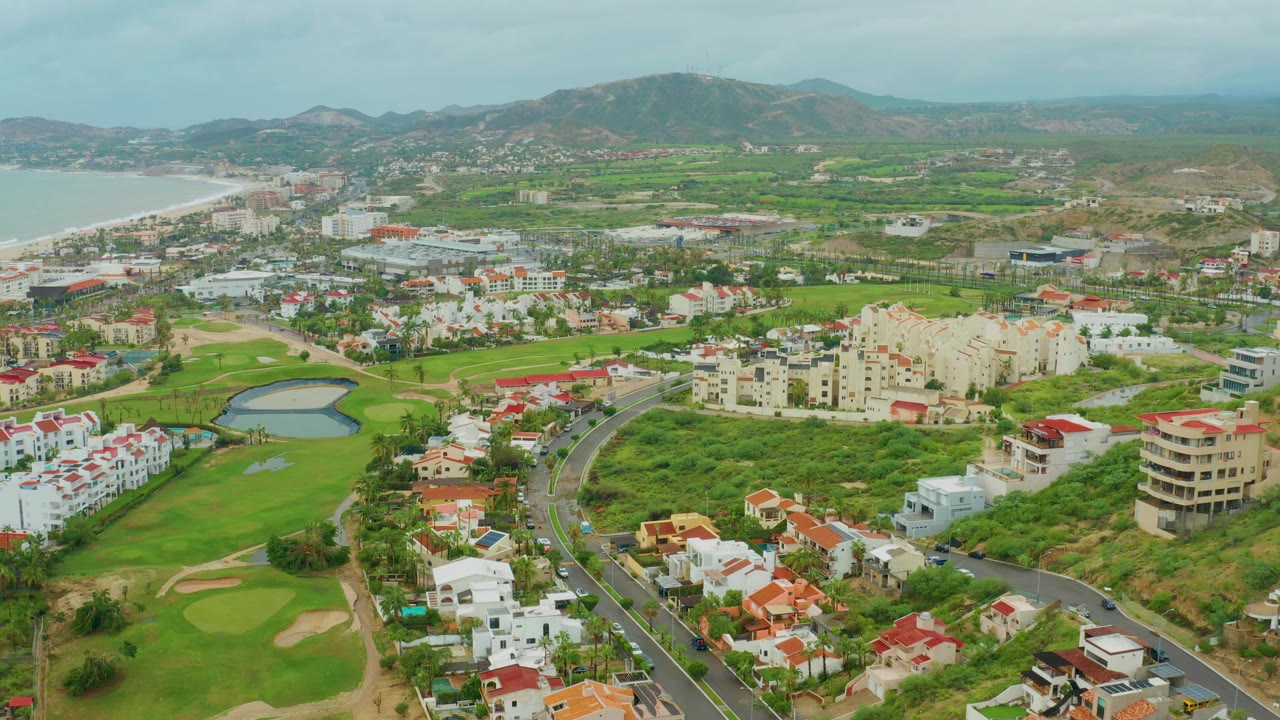 Aerial frame in Baja California Sur, Mexico, showing a curving road with vehicles, green golf fairways and pond, residential buildings, coastline at left, distant hills, and no visible people.