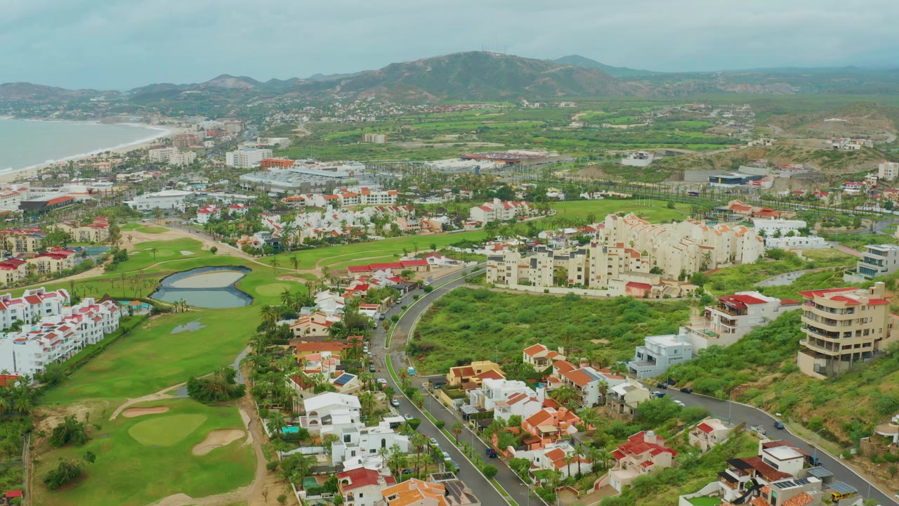 Drone-height view in Baja California Sur, Mexico, with cars on the winding road, bright golf greens and sand traps in the foreground, clustered white buildings, a coastal bay, hazy hills, and no visible people.