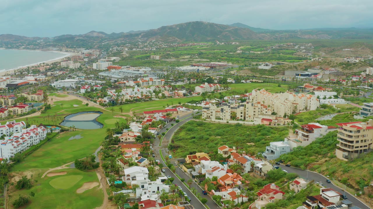 Aerial frame over Baja California Sur, Mexico, showing the pond and golf course more centrally, cars along the curved road, low-rise homes and larger beige buildings, coast to the left, hills beyond, and no visible people.