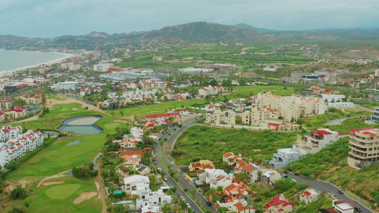 Aerial coastal town view with golf course, curved road, residential buildings, and hills.
