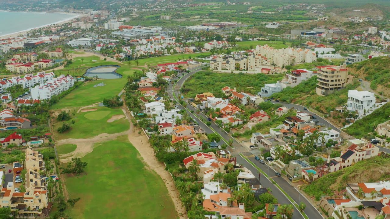 Aerial frame from San Jose Del Cabo BCS Mexico showing the golf course stretching up the center, white residential buildings and red roofs around it, a curving road with small cars, a pond near the upper middle, beach water at left, and no visible people.
