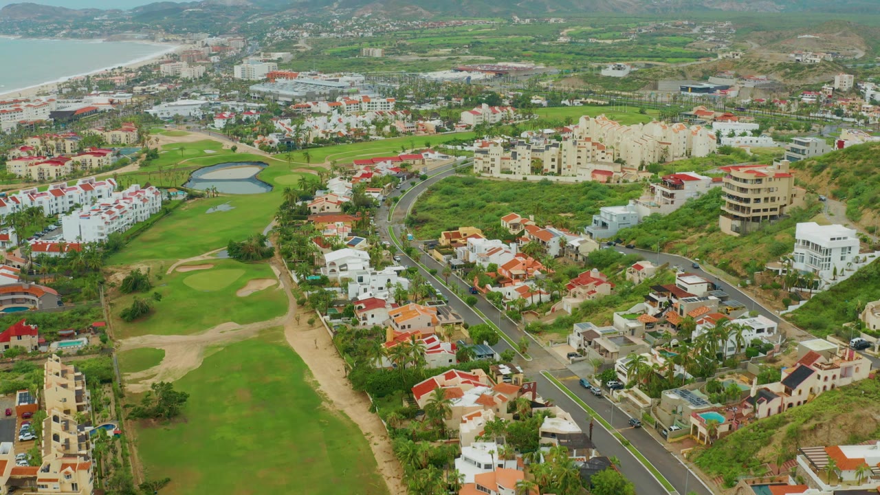 San Jose Del Cabo BCS Mexico aerial view with sand traps and fairways at left-center, villas and apartment blocks to the right, a dark road running diagonally through the housing, coastal water in the upper left, distant hills, and no visible people.