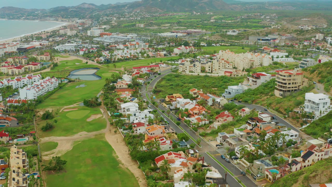 Aerial neighborhood view with golf greens, pond, red roofs, beach water, and mountain ridges.