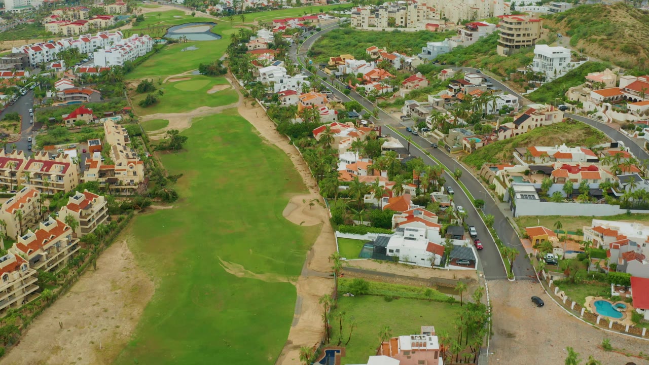 Drone-height view in San Jose del Cabo BCS Mexico with the fairway stretching through the left side, a road and red-roofed homes to the right, hillside buildings beyond, and no visible people.