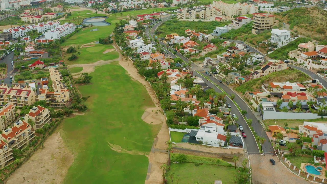 Aerial frame over San Jose del Cabo BCS Mexico showing a broad green fairway, sandy rough, red and white residential roofs, a black road with small cars, hillside vegetation, and no visible people.