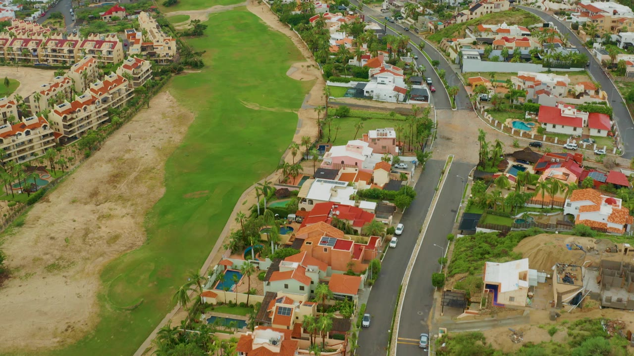 Aerial view in San Jose del Cabo, Baja California Sur, showing a green golf fairway beside red-roofed homes, a curving road, palms, and no visible people.