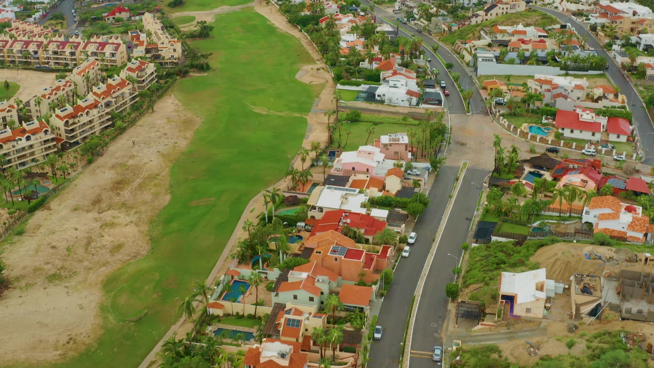 Aerial frame in San Jose del Cabo, Baja California Sur, with the golf course running along the left side, houses and a road on the right, small vehicles, and no visible people.