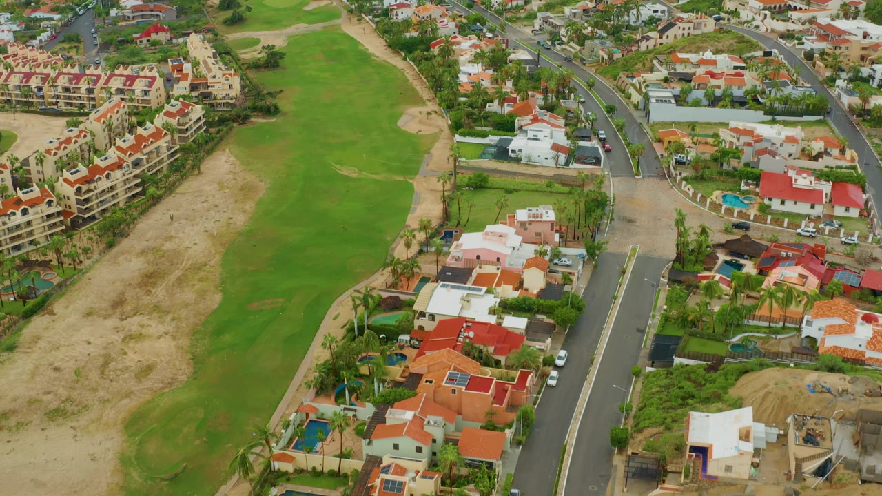 San Jose del Cabo aerial view with a wide green fairway, tan rough and bunkers, red and white residential roofs, a dark paved road, and no visible people.