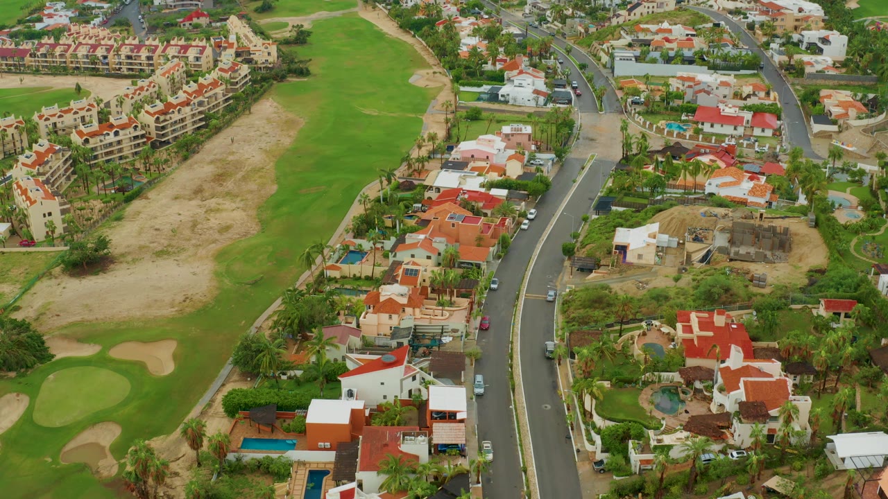 Drone-height view in San Jose del Cabo, Baja California Sur, Mexico, showing red and white residences, backyard pools, a curving road with cars, green golf areas, sandy patches, and no visible people.