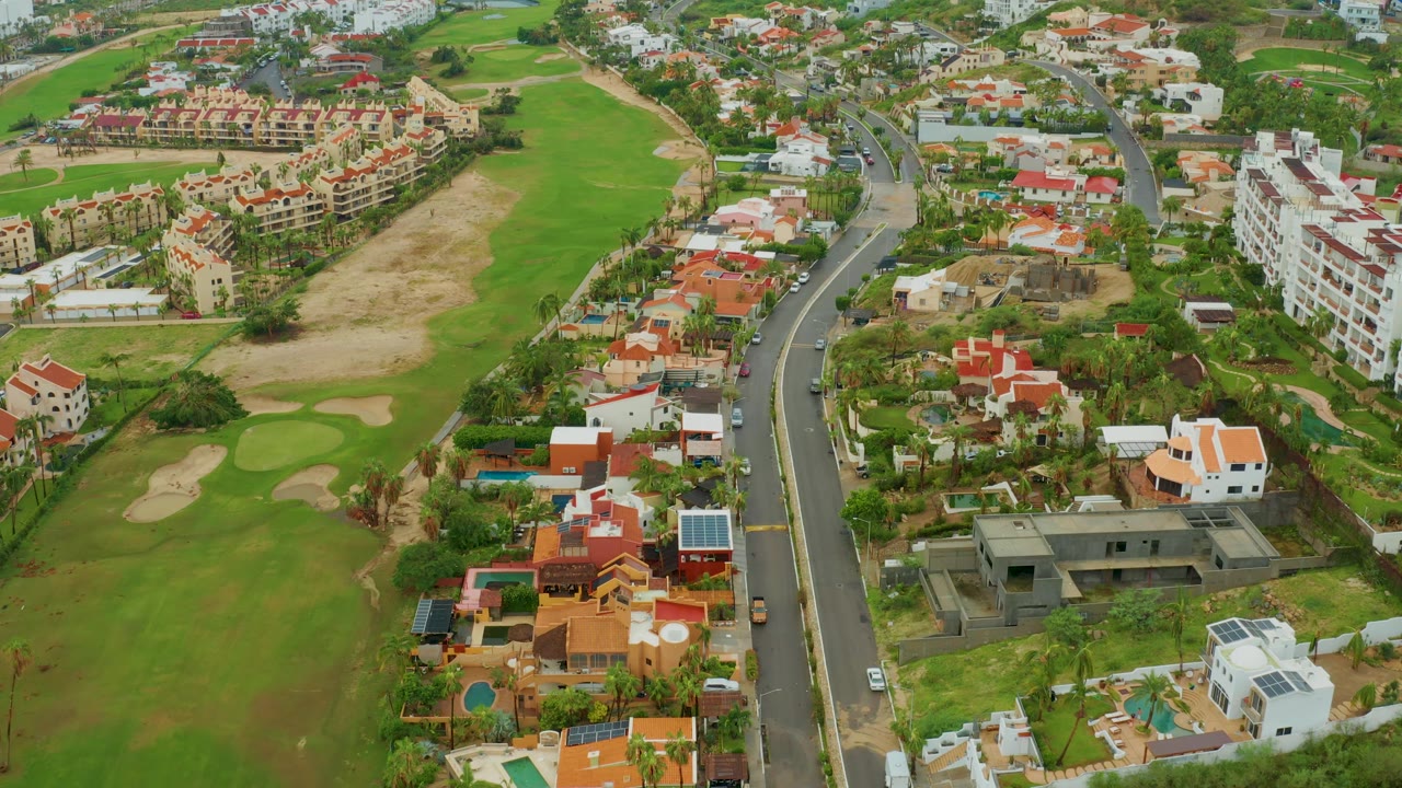 Aerial view in San Jose del Cabo with the curved road more centered, houses on both sides, golf course greens and bunkers to the left, and no visible people.