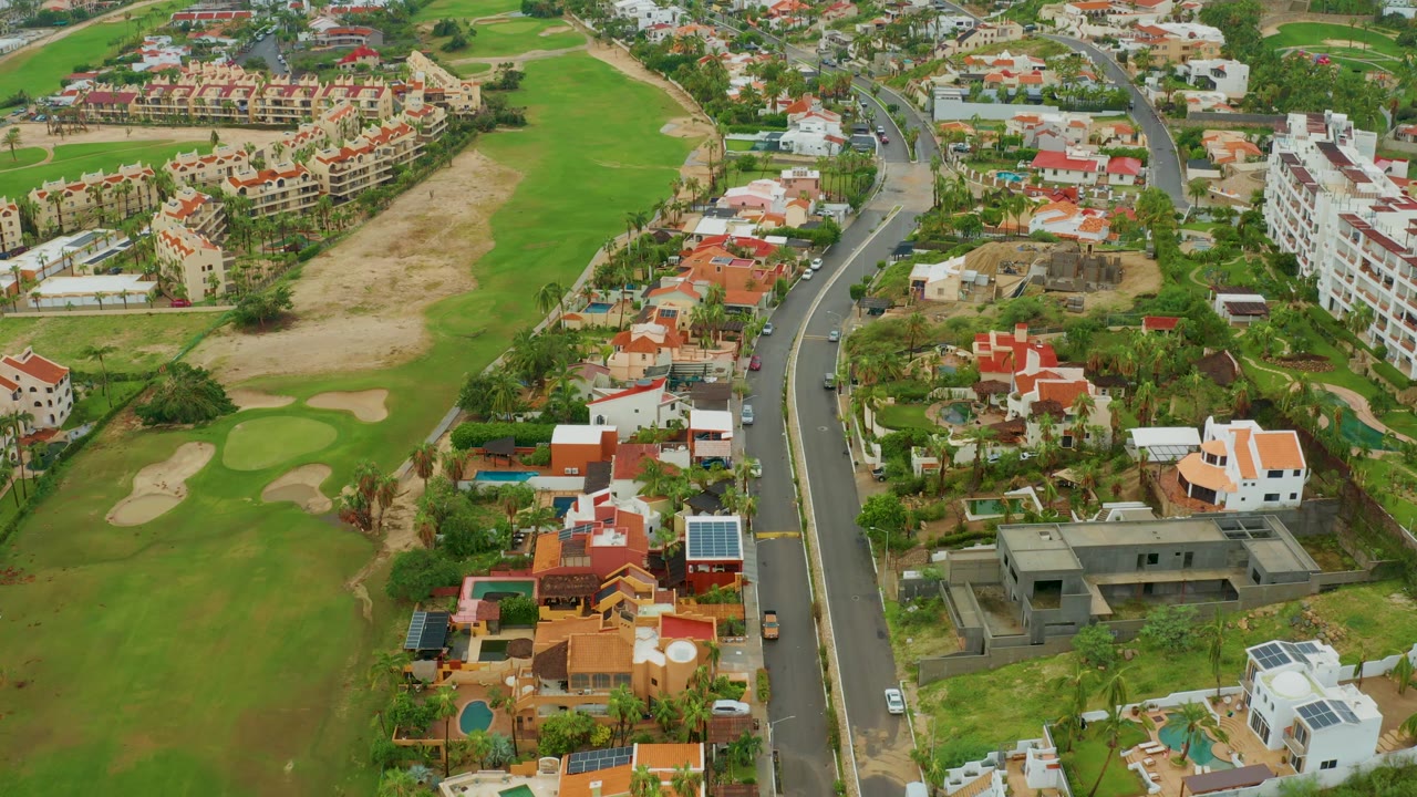 San Jose del Cabo aerial frame showing apartment blocks near the top edge, clustered red-roofed homes, a winding road, golf course bunkers, and no visible people.