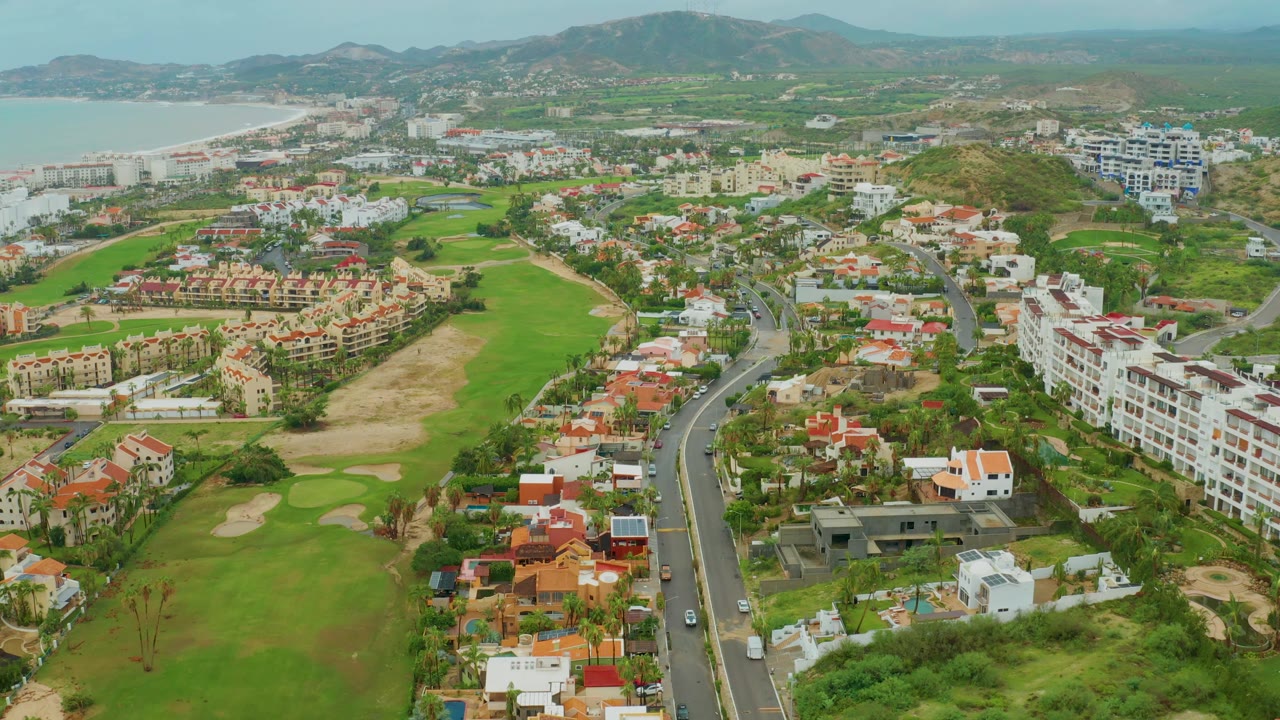 Aerial frame in San Jose del Cabo looking across a golf course and residential blocks toward the coastline, with a central road, small vehicles, white buildings, red roofs, and no visible people.