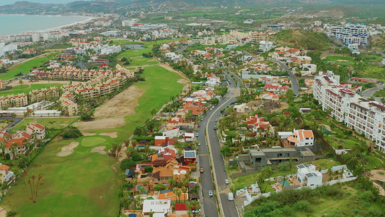 Aerial neighborhood view with golf course, road, red roofs, white buildings, and ocean.